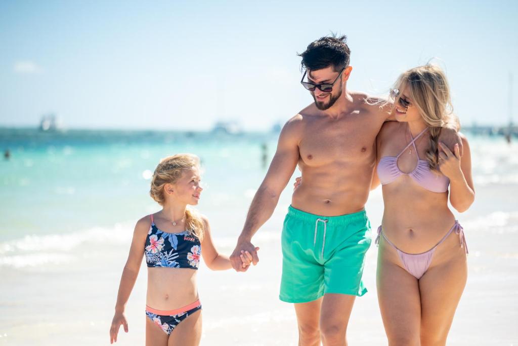 Ģimene pastaigā pa pludmali, kur bērns tur rokās tēvu un māti, kuri ir ģērbti peldkostīmos. Skats uz saulainu dienu un jūras viļņiem.