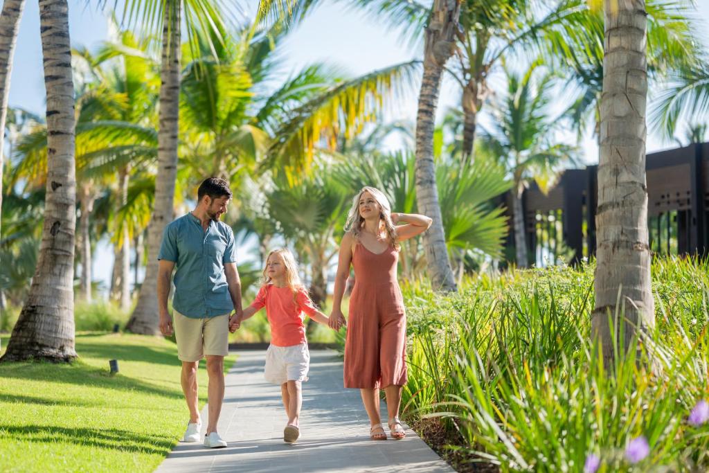 Ģimene pastaigā pa saulainu trotuāru, ko ieskauj zaļi palmu koki un ziedi, radot atpūtas un laimes atmosfēru.