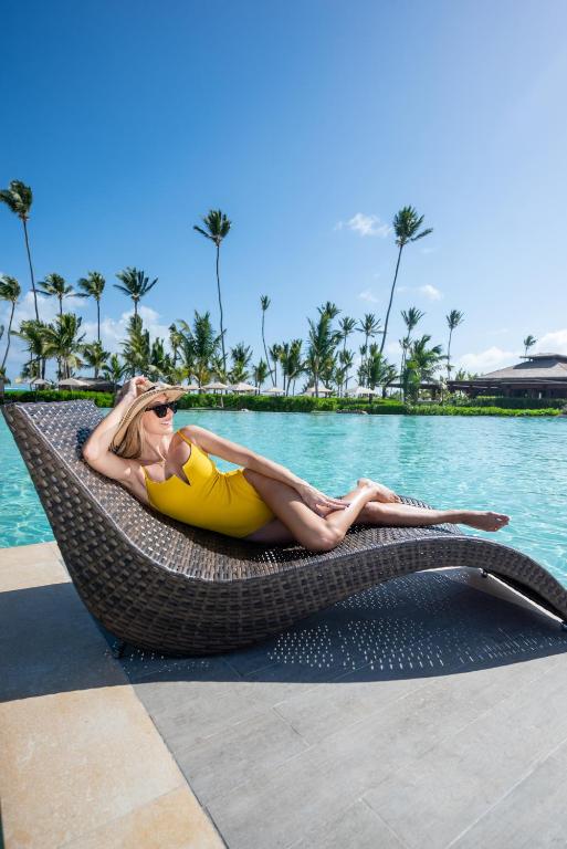 Saulainā pludmales ainava ar sievieti dzeltenā peldkostīmā, kas atpūšas uz modernās sauļošanās krēsla pie baseina, ieskauta palmu un zaļo zālienu.