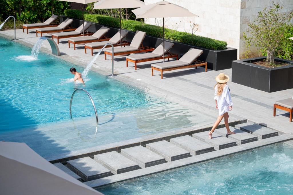 Peldbaseins ar pludmales krēsliem un saulessargiem, kurā redzama sieviete ar cepuri, kas pastaigājas pa akmens celiņu, kamēr vīrietis peld ūdenī.