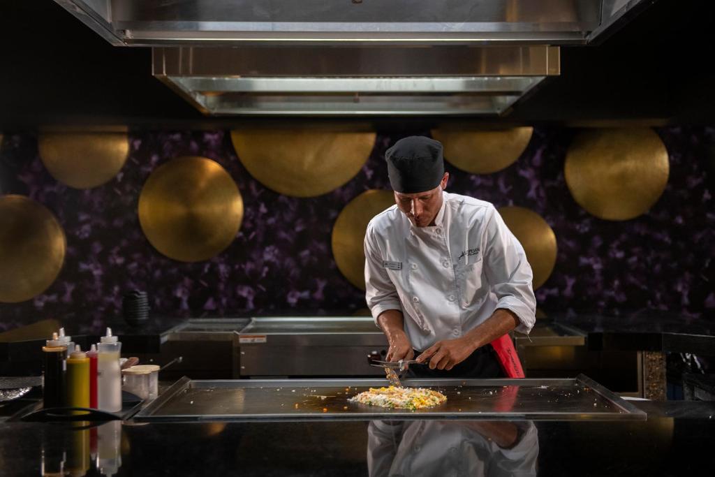 Šefpavārs gatavo ēdienu uz plīts, izmantojot svaigus dārzeņus un rīsus. Fons ir ar zeltainām dekorācijām un modernu dizainu.