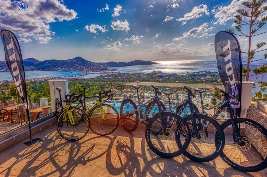 Kalnu un ceļu velosipēdi novietoti terasē ar panorāmas skatu uz jūru un kalniem. Fons izceļ skaistu saulrietu un mierīgu ainavu.