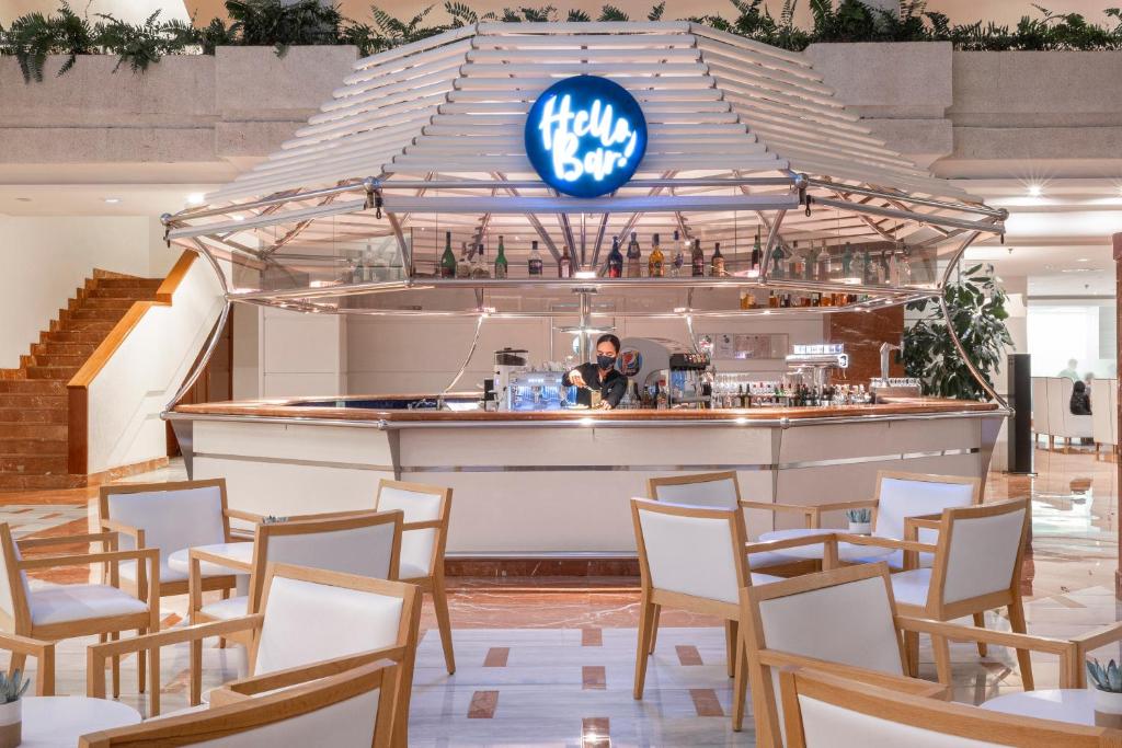 Mūsdienīgs bārs ar stilīgu dizainu un gaišu apgaismojumu, kur bārmenis sagatavo dzērienus. Apkārt atrodas komfortabli krēsli un zaļumi, kas rada patīkamu atmosfēru.