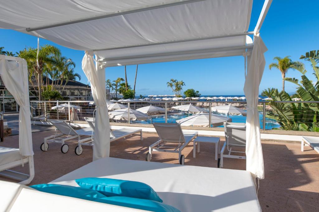 Atpūtas zonas skats ar sauļošanās krēsliem pie baseina un palmām, kas rada relaksējošu atmosfēru. Skaidra debess un zils jūras ūdens papildina brīvdienu sajūtu.