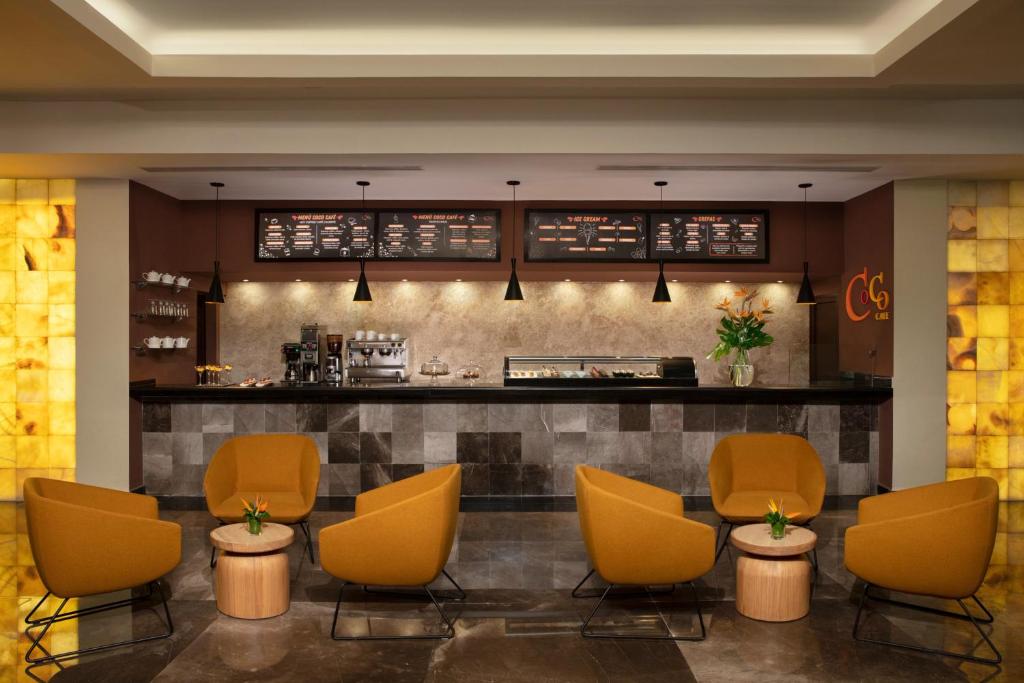 Mūsdienīgs kafijas veikals ar stilīgu interjeru, kas ietver oranžas krēslus un melnu leti. Uz sienas redzama ēdienkarte, un telpā ir ziedi, radot patīkamu atmosfēru.