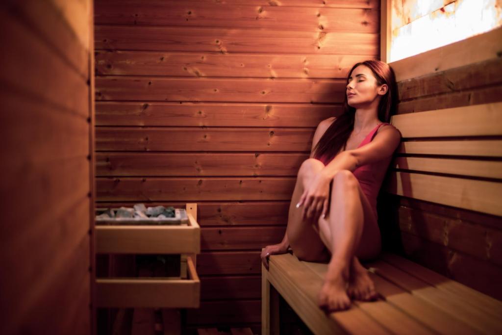 Sieviete relaksējas koka pirtī, sēžot uz soli un izbaudot siltumu. Pirts interjers ir izgatavots no koka, radot mierīgu atmosfēru.