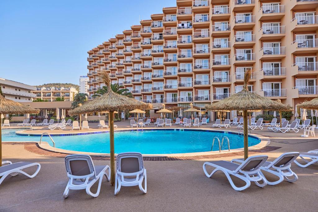 Peldbaseins ar sauļošanās krēsliem un palmu lapu nojumiem viesnīcas pagalmā, kas piedāvā relaksējošu atmosfēru un komfortu. Viesnīcas ēka ar balkoniem atrodas fona.