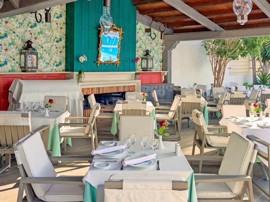 Gaiša un eleganta restorāna iekārtojums ar kokvilnas galdautiem, ziediem uz galdiem un krāsainu fonu. Vides elementi, piemēram, lustras un spoguļi, piešķir atmosfēru.