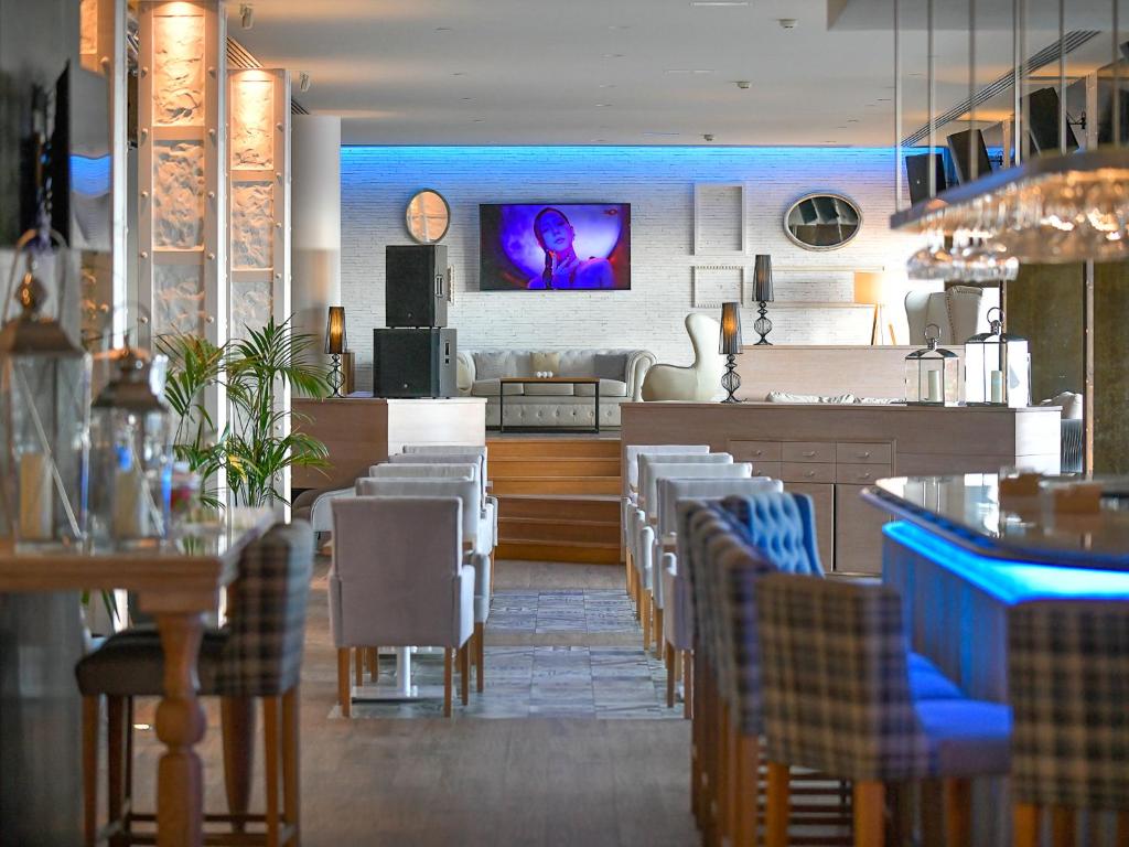 Mūsdienīgs restorāns ar elegantu interjeru, kurā ir redzams televizors, atpūtas zona un modernas mēbeles. Apgaismojums rada patīkamu atmosfēru.