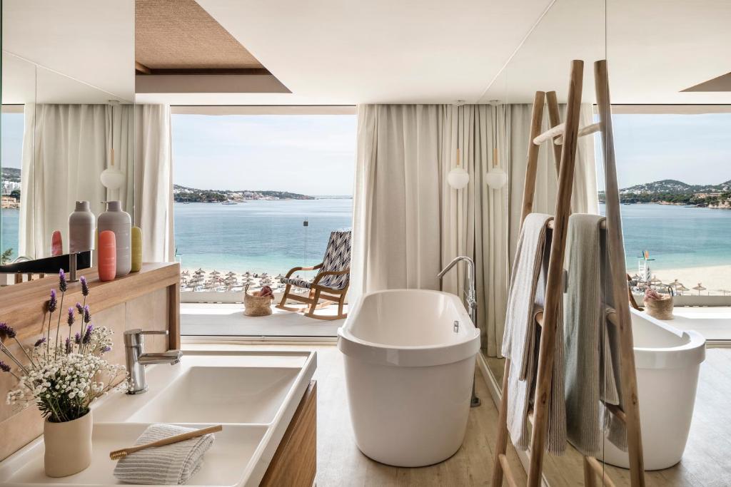 Mūsdienīgs vannas istabas interjers ar skatu uz jūru, kurā redzama brīnišķīga vanna, vannas izlietne un dabīgās gaismas pieplūdums. Telpa ir eleganta un gaiša, radot relaksējošu atmosfēru.