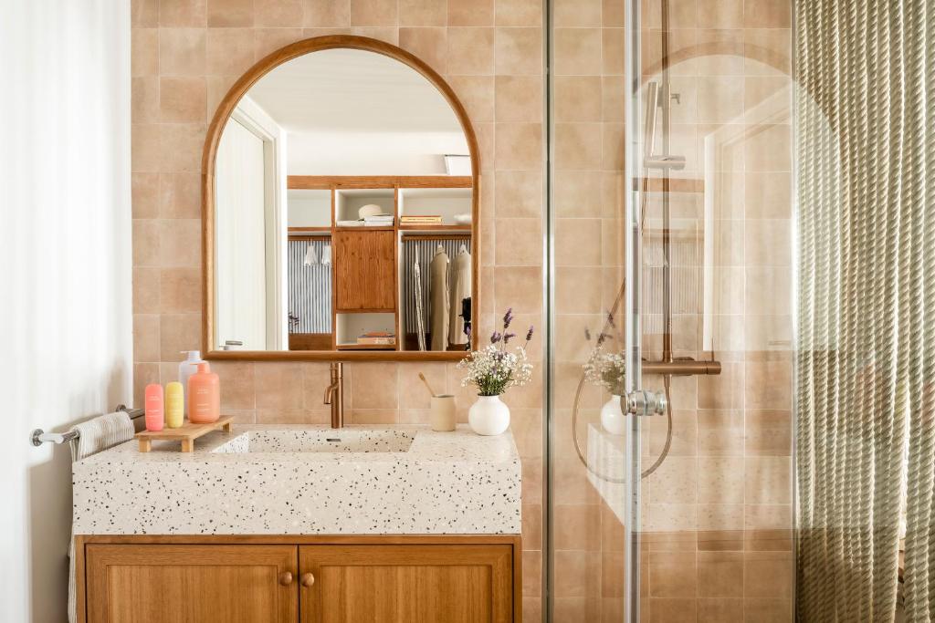 Mūsdienīgs vannas istabas interjers ar apaļu spoguli, terakotas krāsas flīzēm un marmora izskata izlietni. Uz plaukta atrodas dažādas ķermeņa kopšanas līdzekļu pudeles un ziedu kompozīcija.