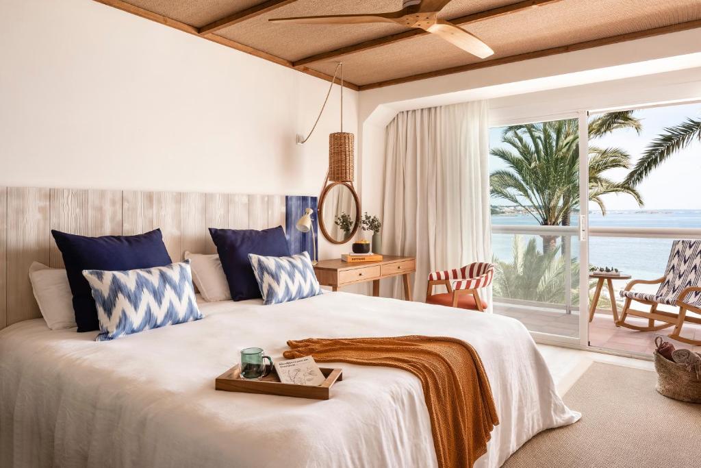 Gaišs un elegants guļamistabas interjers ar skatu uz jūru, iekārtots ar zilu un balto tekstilu, koka mēbelēm un dabīgu apgaismojumu.