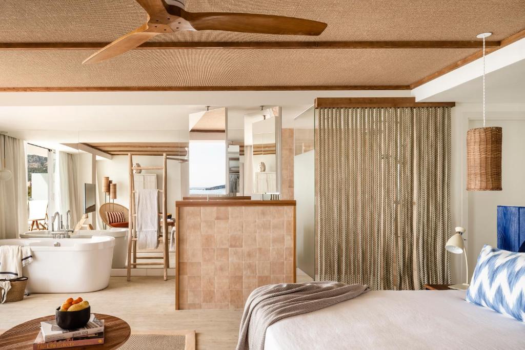 Moderns guļamistabas interjers ar atvērtu plānojumu, kurā redzams vannas istabas stūris, liels, balts vanna, un dabīgi materiāli dekorēti ar koka elementiem. Gaisma plūst cauri logiem, radot relaksējošu atmosfēru.