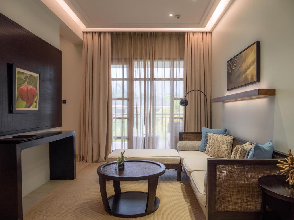 Mūsdienīgs viesnīcas numurs ar elegantu mēbelējumu, gaišām aizkariem un dabīgu apgaismojumu. Interjerā ir ērta atpūtas zona ar dīvānu un galdiņu, radot relaksējošu atmosfēru.