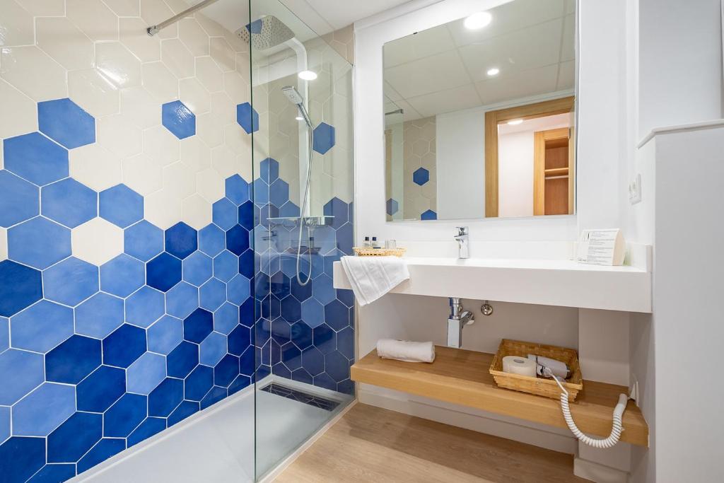 Mūsdienīga vannas istaba ar ģeometrisku zilo šūnu flīžu dizainu un stikla dušas kabīni. Uz izlietnes atrodas dvielis un higiēnas piederumi.