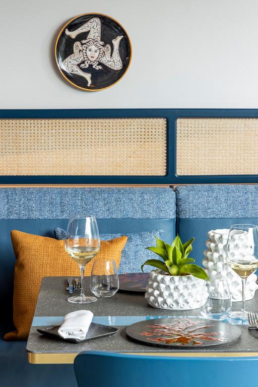 Mūsdienīgs restorāna interjers ar zilu dīvānu, dekoratīvu trauku pie sienas un galdu, kas klāts ar vīna glāzēm un augu. Stilīgas detaļas un krāsainas akcenti rada viesmīlīgu atmosfēru.