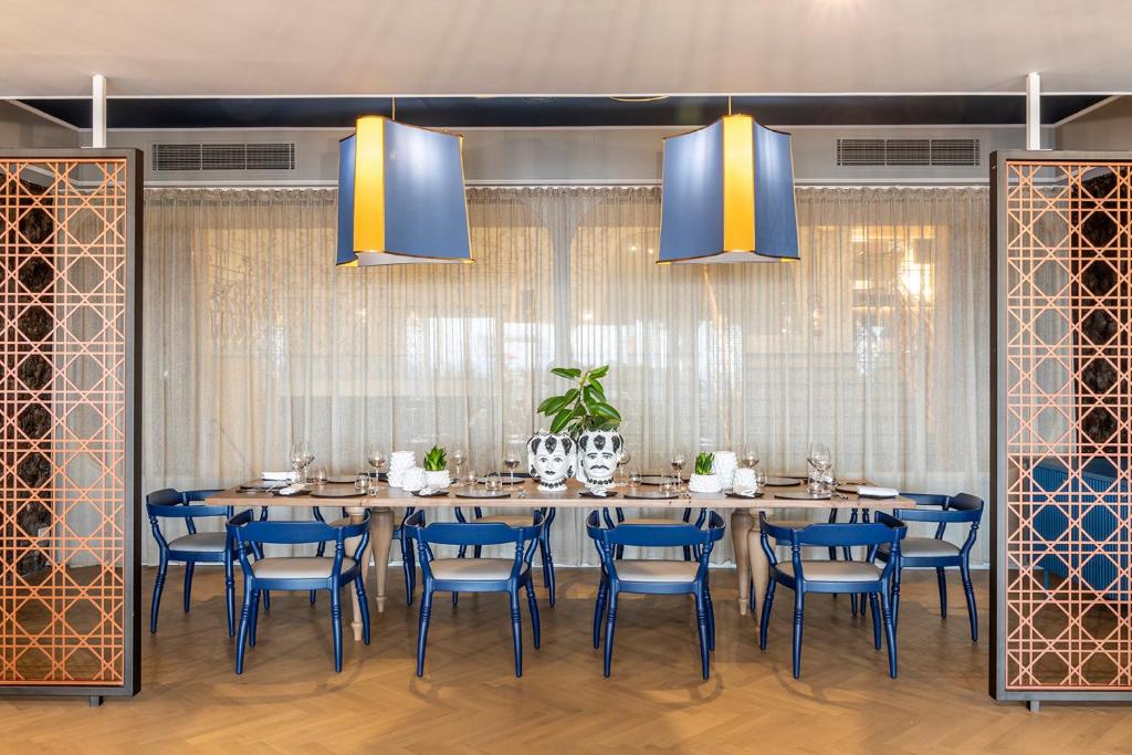 Mūsdienīgs restorāna interjers ar ilgu galdu, zilām krēslām un dekoratīvām lampām, kas piedāvā siltu atmosfēru. Aiz galda redzams augu kompozīcija un elegantas galda uzkodas.