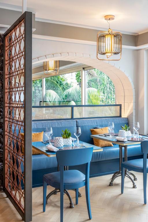 Mūsdienīgs restorāna interjers ar zilu dīvānu, elegantiem galdiem un modernām lampām, kas rada patīkamu atmosfēru. Logi atver skatu uz zaļo dārzu, piešķirot telpai gaisīgu noskaņu.