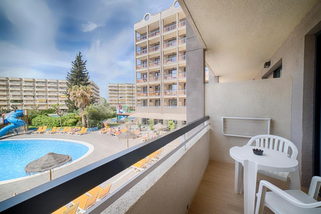 Balcony view of a hotel overlooking a swimming pool and sun loungers, surrounded by greenery and a water slide. Perfect for a relaxing holiday.
