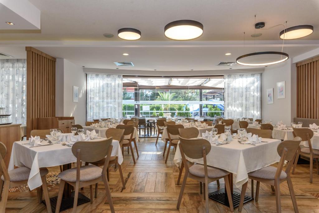 Mūsdienīgs restorāns ar eleganti klātiem galdiem un plašu logu, kas piedāvā skatu uz zaļo dārzu. Gaisma un koka elementi rada siltu un viesmīlīgu atmosfēru.