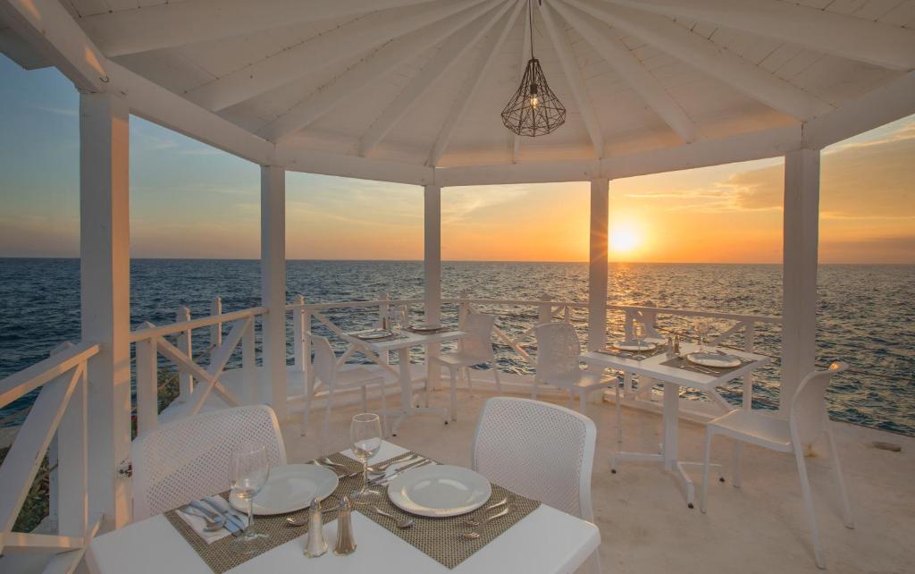 Romantiska pludmales restorāna terase ar skatu uz saulrietu virs okeāna, eleganti galdi un krēsli.