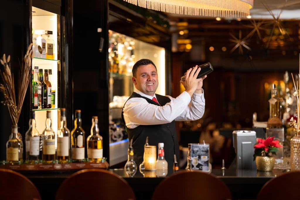 Bārmene, kas sajauc kokteiļus, stāv aiz elegantā bāra ar dažādiem dzērieniem un rotājumiem. Interjers ir grezs un viesmīlīgs, radot patīkamu atmosfēru.