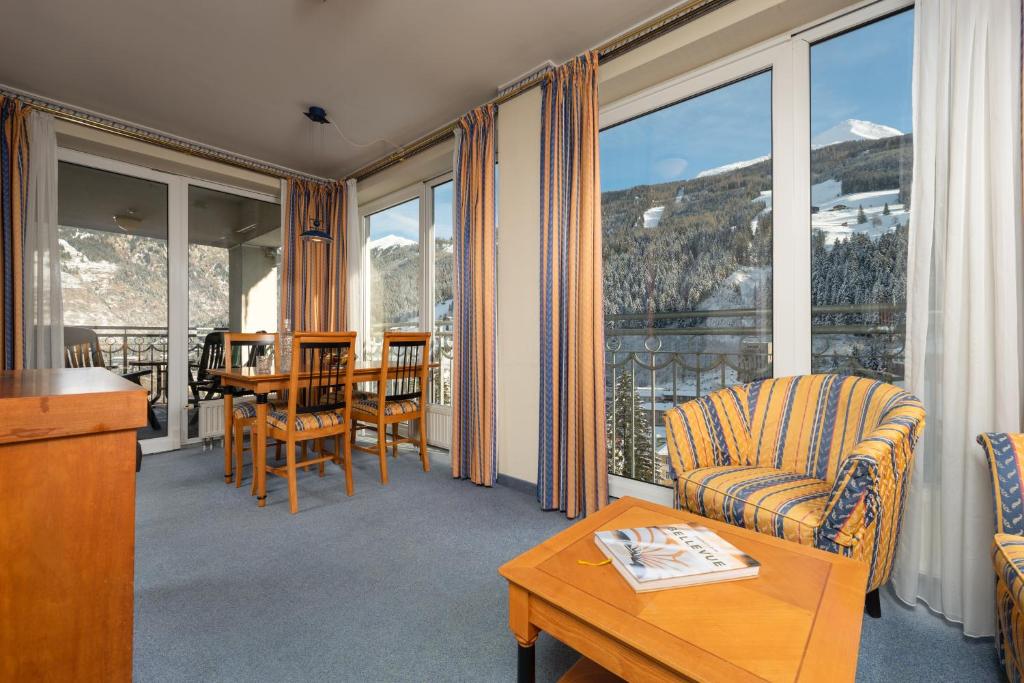 Mūsdienīgs interjers ar viesistabu, kurā ir koka galds un krēsli, dīvāns ar svītrām, un plašas logu durvis, kas paver skatu uz kalnu ainavu.