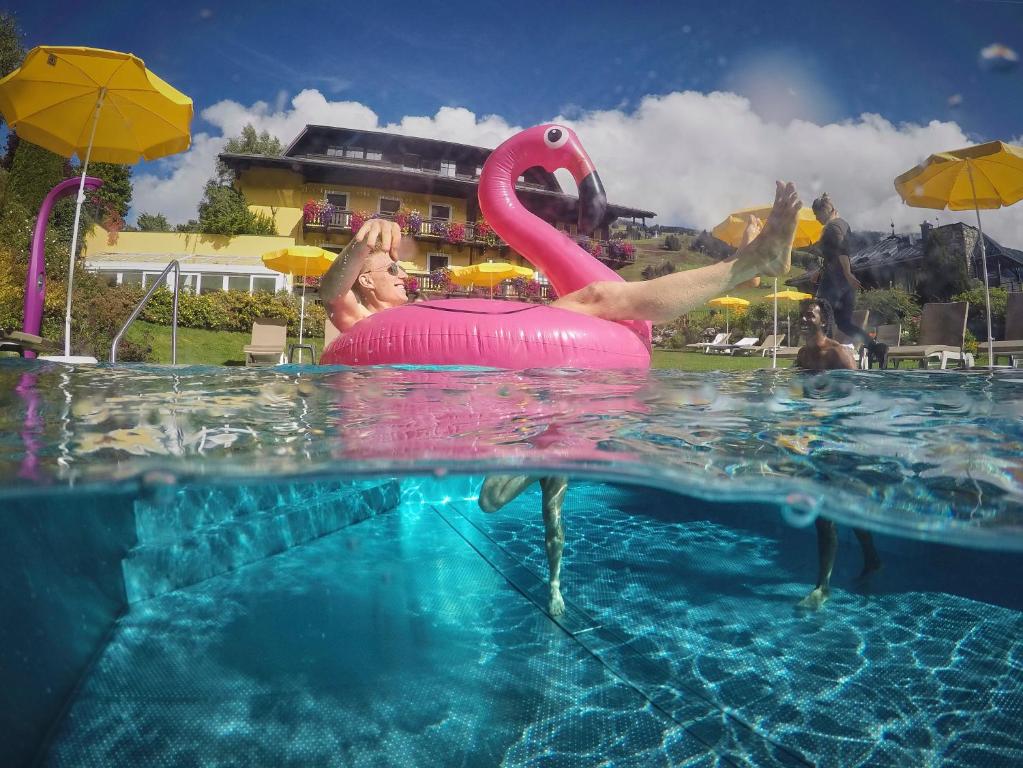 Persona, kas peldē rozā pīlītes formā, atpūšas baseinā ar saulessargiem un viesnīcu fonā. Vasaras brīvdienu atmosfēra.