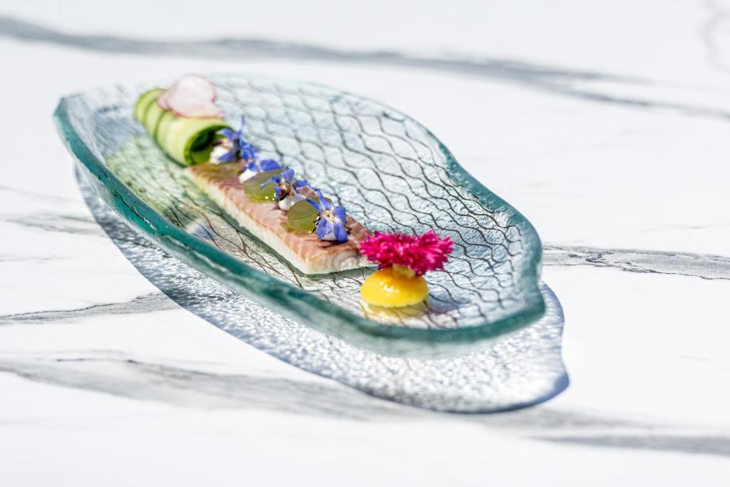 Elegants ēdiens uz stikla šķīvja, kas satur zivi, dārzeņu rotājumu un ēdamus ziedus, piedāvā estētisku un gastronomisku pieredzi.