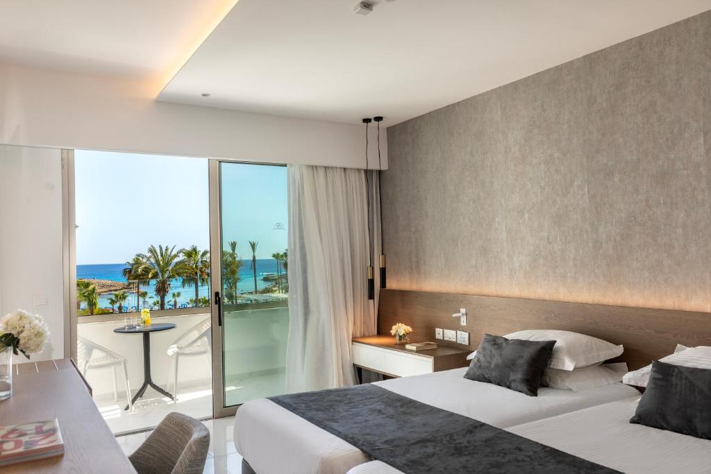 Mūsdienīgs viesnīcas numurs ar skatu uz jūru un palmām, iekārtots ar gultām un elegantu interjeru. Balkons ar mazu galdu un krēsliem piedāvā relaksējošu atmosfēru.