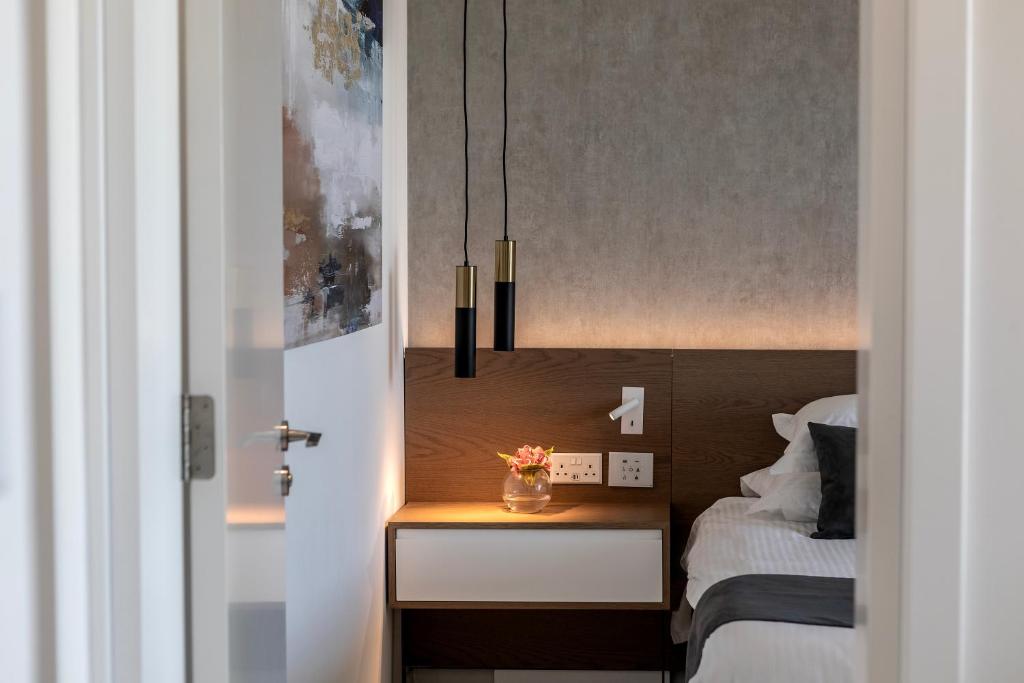 Mūsdienīga guļamistabas interjera skats ar gaismas ķermeņiem un elegantu naktsgaldiņu, kas rotāts ar ziedu pušķi.