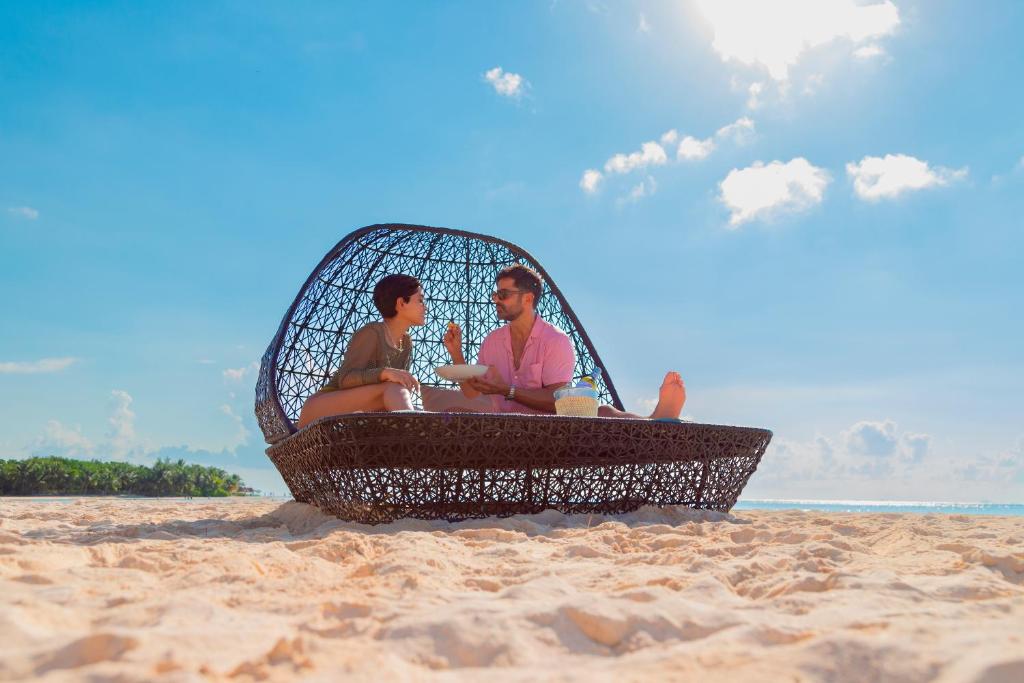 Pāris atpūšas pludmalē, sēžot modernā režģveida krēslā, baudot dzērienus un sauli. Fons ir skaidrs debesis un jūra, radot relaksējošu atmosfēru.