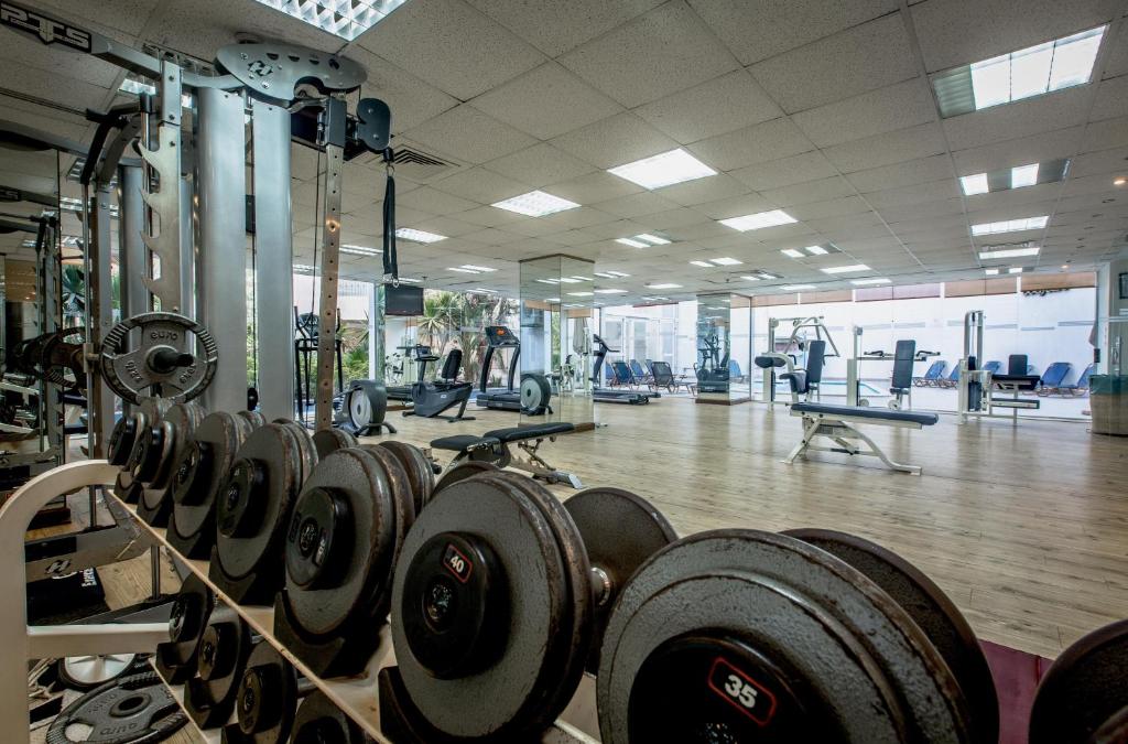 Moderns sporta zāles iekārtojums ar svaru stieņiem priekšplānā un trenažieriem fonā. Gaismas un telpas dizains rada iedvesmojošu vidi aktīvam dzīvesveidam.