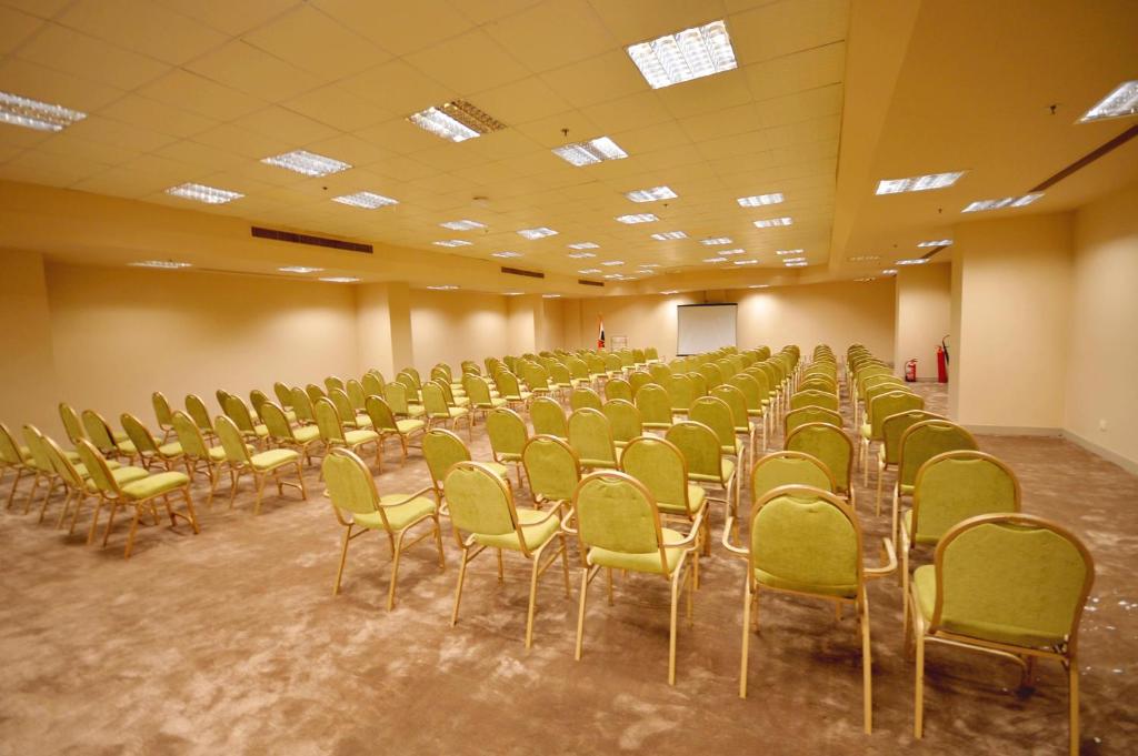 Konferenču zāle ar zaļām krēslām, sakārtotām rindās, un projekcijas ekrānu priekšā. Telpa ir gaiša un plaša, piemērota pasākumiem un sanāksmēm.