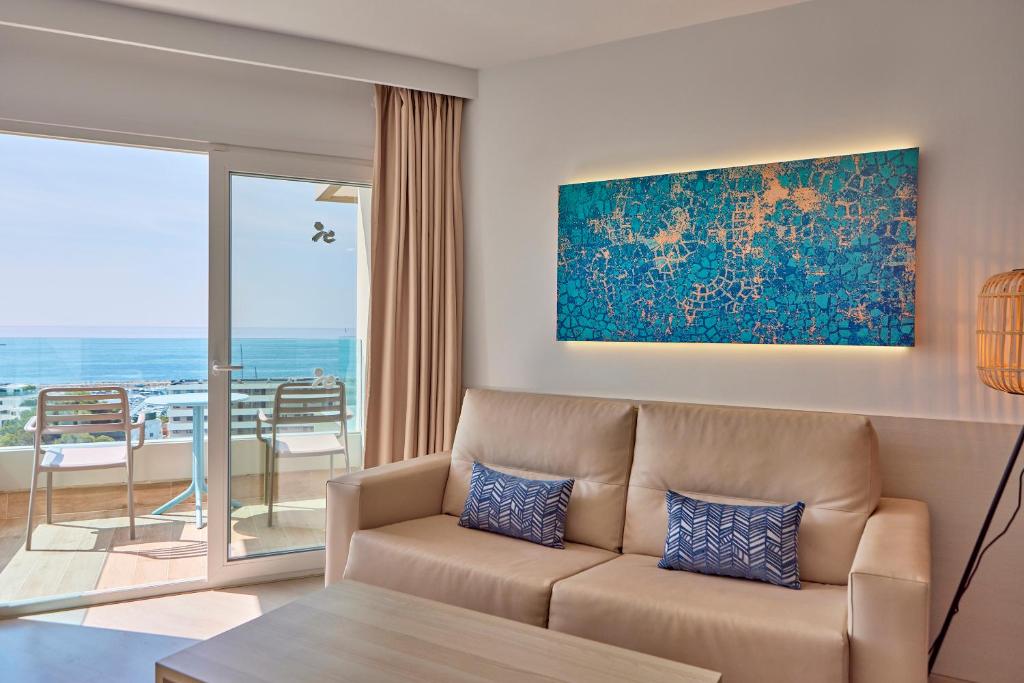 Mūsdienīgs viesistabas interjers ar jūras skatu, gaišām krāsām un dekoratīvu mākslas darbu. Sofā ir divas spilvendizaines ar ģeometrisku rakstu, bet blakus ir balkons ar krēsliem.