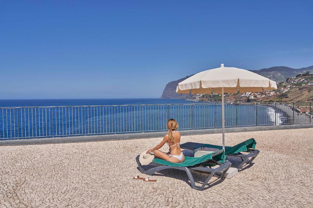 Saulaina pludmales ainava ar cilvēku, kas atpūšas uz sauļošanās krēsla, skatoties uz jūru un kalnu ainavu. Ideāla vieta relaksācijai un atpūtai.