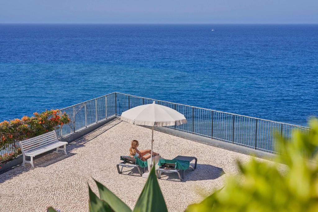 Atpūtas zona ar sauļošanās krēsliem un lietussargu pie jūras, kur sieviete relaksējas un izbauda skatu uz zilo ūdeni.