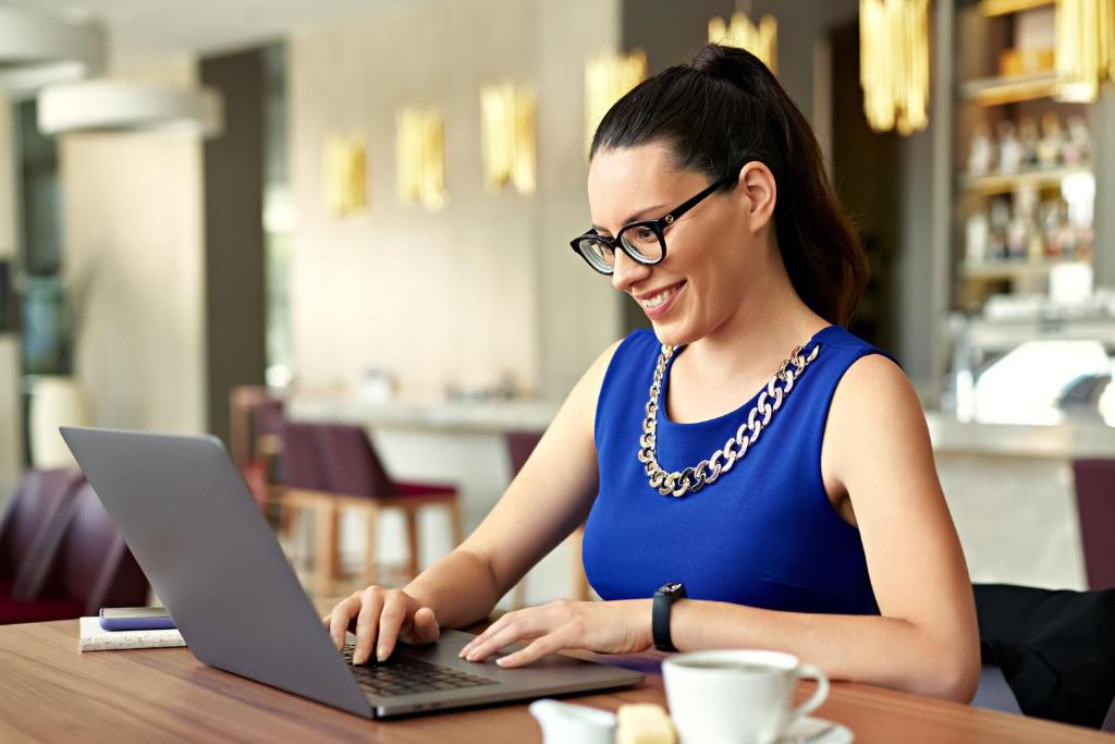 Jauna sieviete strādā pie datora kafejnīcā, valkājot zilu kleitu un brilles. Viņa smaida, koncentrējoties uz ekrānu, kam blakus ir kafijas tasīte un piezīmju bloks.