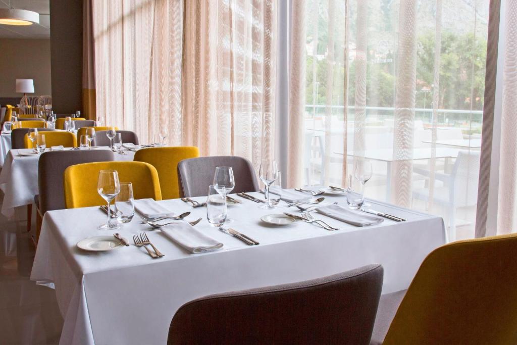 Mūsdienīgs restorāns ar eleganti klātām galdiem, kas aprīkoti ar baltiem galdautiem un krāšņām krēsliem. Labi apgaismots interjers ar lieliem logiem un mīkstām aizkariem, kas nodrošina patīkamu atmosfēru.