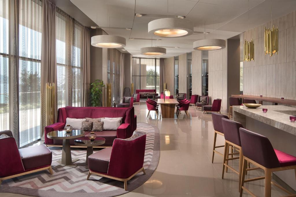 Moderns viesnīcas interjers ar elegantiem sarkaniem dīvāniem un gaišu apgaismojumu, radot komfortablu un stilīgu atmosfēru. Labi organizēta telpa ar galdu un krēsliem, ideāli piemērota atpūtai vai sanāksmēm.