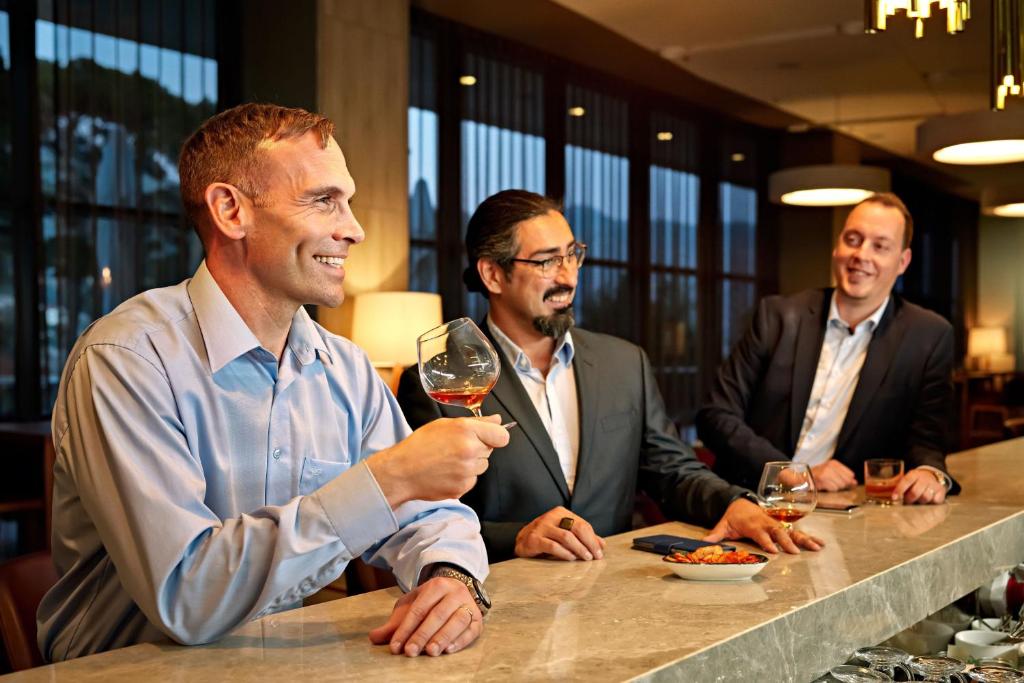 Trīs vīrieši, ģērbušies uzvalkos, izbauda dzērienus un uzkodas modernā bārā, smejoties un sarunājoties. Interjers ir stilīgs un elegants.