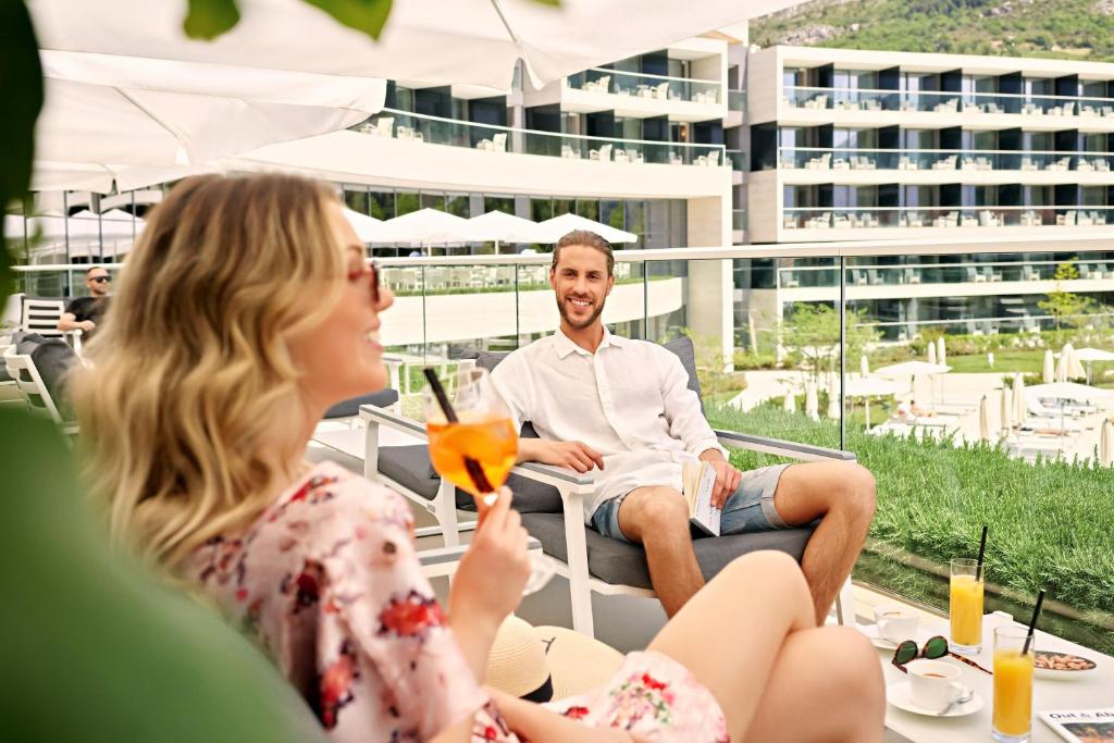 Skaisti cilvēki relaksējas pie baseina, baudot dzērienus un saules gaismu, apņemti ar modernu viesnīcas fonu.