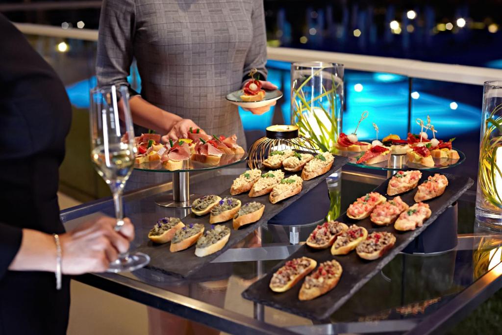 Elegants pasākums ar dažādām uzkodām uz galda, tostarp maizītēm ar dažādām piedevām un šampanieti rokās. Gaisma un ūdens fons piešķir atmosfēru.