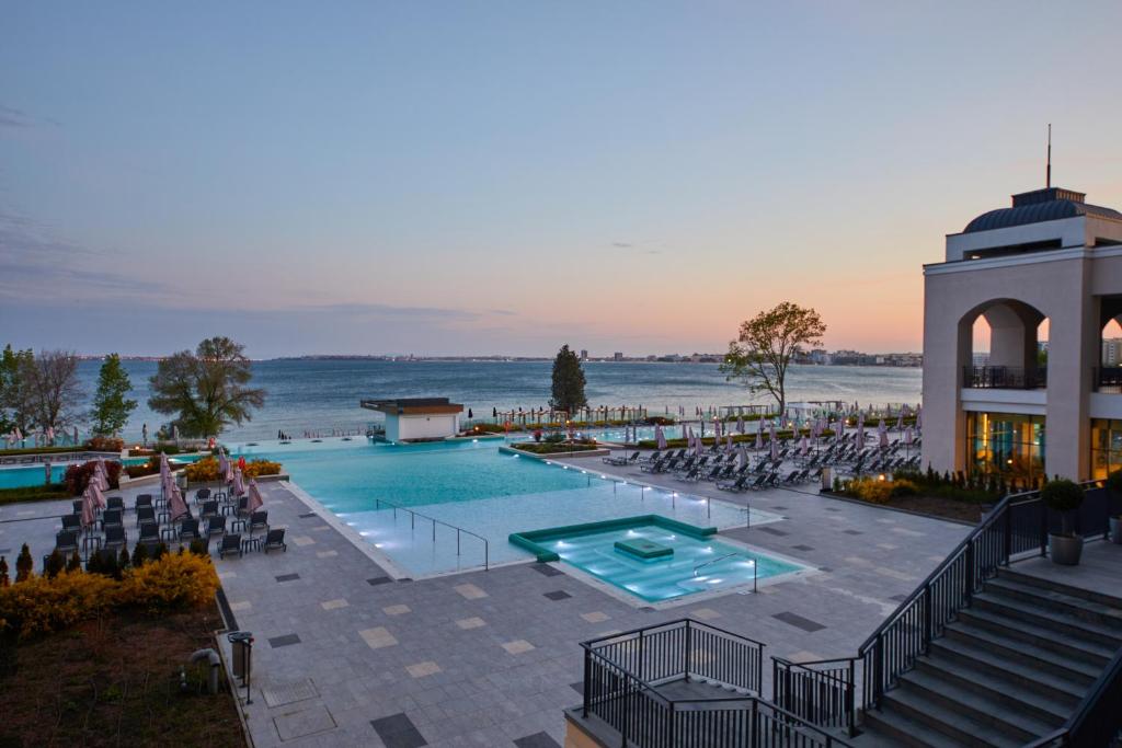 Peldbaseins ar skatu uz ūdenskrātuvi un saulrietu, apzaļumots apkārtējais laukums ar atpūtas krēsliem un terasi.