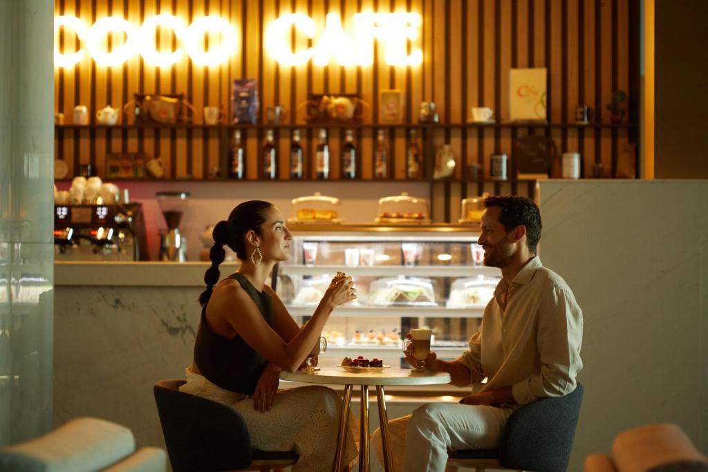 Divi cilvēki sēž kafejnīcā, baudot dzērienus un desertus. Kafejnīcas interjers ir moderns, ar spilgtām gaismām un izsmalcinātām detaļām.