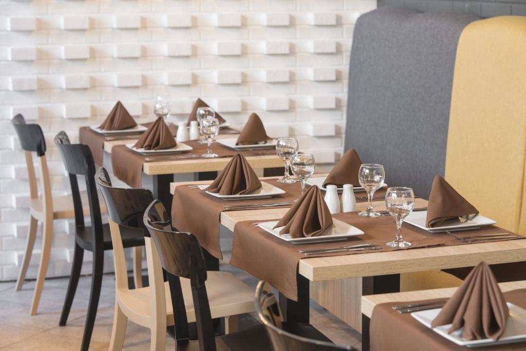 Elegantā restorāna iestatījums ar galdautiem un salvetēm brūnā krāsā, glāzēm un ēdiena traukiem. Fons veidots ar teksturētu baltu sienu un krāsainām atzveltnēm.