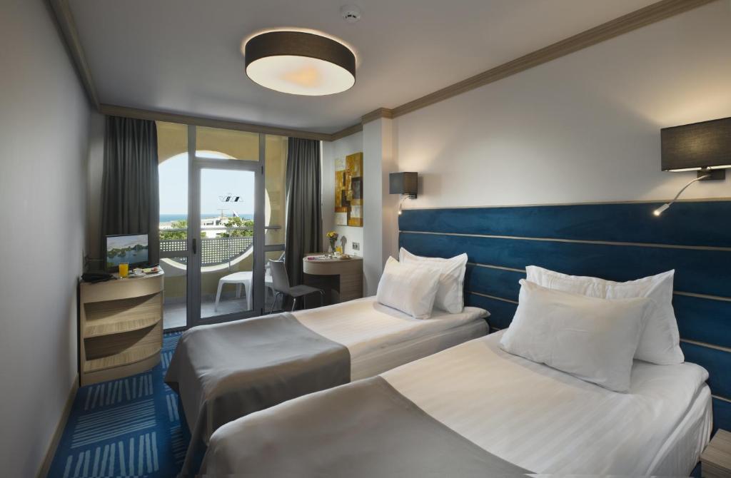 Mūsdienīgs viesnīcas numurs ar divām gultām, koka mēbelēm un skatu uz jūru. Numurā ir televizors, galdiņš un balkons ar durvīm, kas ved uz āru.