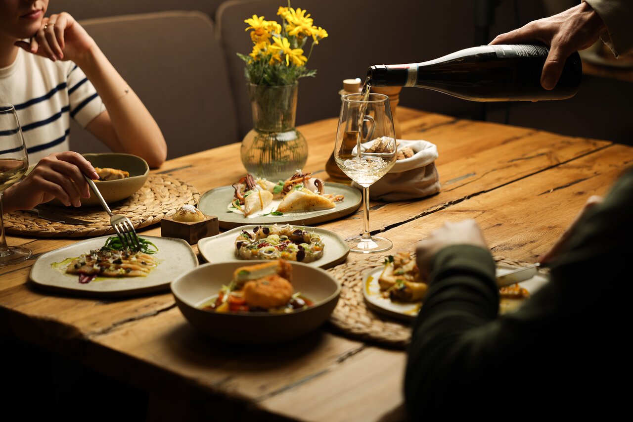 Elegantā vakariņu aina ar dažādiem ēdieniem un vīnu, kas tiek pasniegts pie koka galda, rotāta ar ziediem. Siltā atmosfērā redzamas divas personas, kas baudīja maltīti kopā.