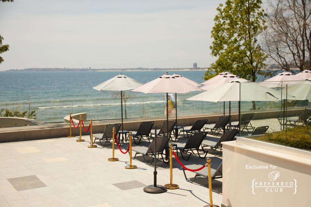 Atpūtas zona pie ezera ar sauļošanās krēsliem un rozā saulessargiem, kas nodrošina ēnu. Skats uz ūdeni un apkārtējiem kokiem rada relaksējošu atmosfēru.