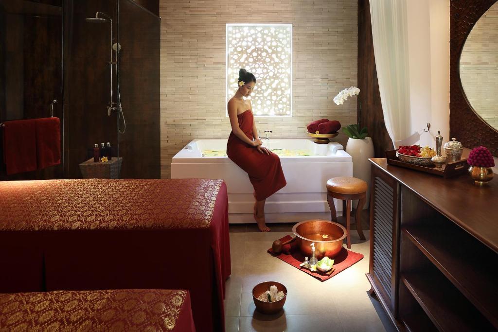 Elegants spa interjers ar relaksējošu atmosfēru, kur sieviete sēž pie vannas ar ziediem un aromātiskām sveču gaismām. Sarkanā krāsa un dabīgie materiali rada mierīgu noskaņu.